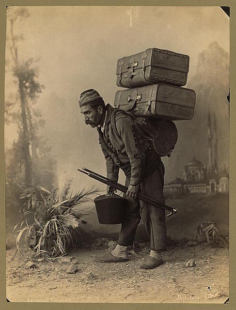 Photo ancienne : un porteur turque, cassé en deux, porte sur son dos de lourds bagages